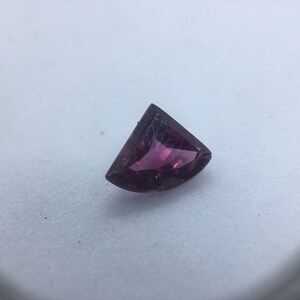Puede incluir: Una piedra preciosa triangular facetada con un tono rosa intenso. La piedra preciosa está tallada en una forma única y tiene un alto nivel de brillo.