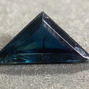 May include: A triangular blue gemstone with a dark blue hue and a slight green tint. The gemstone is cut into a faceted shape and has a smooth, polished surface.