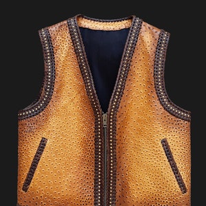 Può includere: Un gilet in pelle marrone chiaro con chiusura a zip e due tasche frontali. Il gilet presenta una superficie strutturata con bordatura intrecciata marrone e dettagli dorati sui bordi. La fodera interna è nera.