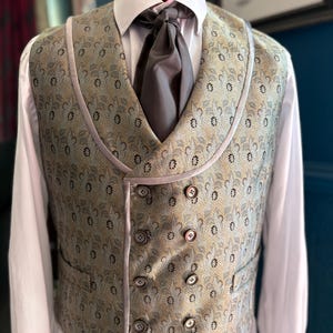 May include: A double-breasted waistcoat featuring a gold and blue leaf pattern, a shawl collar, and eight buttons. The garment is worn over a white collared shirt with a solid grey cravat. The waistcoat has a smooth, light-colored trim along the edges.