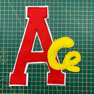 Può includere: Lettera "A" in feltro rosso con bordo bianco e lettera corsiva "c" in feltro giallo con cuciture gialle. Le lettere sono su un tappetino da taglio verde.