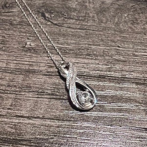 May include: A silver-toned necklace with an infinity symbol pendant. The pendant is adorned with small, clear gemstones and a larger, round gemstone at the center. The necklace chain is delicate and thin.