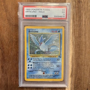 May include: A holographic Pokemon trading card featuring Articuno, a legendary bird Pokemon with blue and white feathers. The card is from the 1999 Fossil set and has a PSA graded score of 5. The card text reads "Freeze Dry" and "Blizzard".