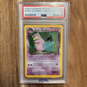 May include: A graded 2000 Pokémon Rocket Dark Slowbro Holo card. The card features a pink Slowbro with a blue shell and the text "Dark Slowbro 60 HP". The card is encased in a protective plastic holder.