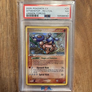 May include: A PSA graded Pokemon card featuring Hitmontop from the Unseen Forces set. The card is a reverse foil and is in near mint condition. The card number is #26 and the grade is 7.