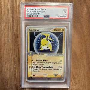 May include: A holographic Pokemon trading card featuring Raichu ex, a yellow electric-type Pokemon with a lightning bolt on its head. The card is from the 2003 Pokemon EX Sandstorm set and has a PSA grading label with a grade of 5. The card's text reads "Raichu ex 100 HP, Stage 1, Dazzle Blast 20, Mega Thunderbolt 120".