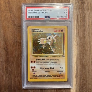 May include: A holographic Hitmonlee Pokemon card from the 1999 Fossil set. The card is in excellent condition and has a PSA grading label. The card features a yellow and brown Hitmonlee with a gold background. The card text reads "Stretch Kick" and "High Jump Kick".