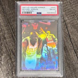 Peut inclure: Une carte de collection Michael Jordan 1991 U.D. Award Winner Holograms, dans un étui de protection. La carte présente des images de Michael Jordan en action, avec le texte "MICHAEL JORDAN SCORING" en bas.