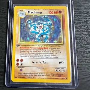 May include: A blue and white Pokemon trading card featuring Machamp, a Stage 2 Pokemon with 100 HP. The card has a blue background with stars and a white border. The card text describes Machamp's Pokemon Power, Strikes Back, and its attack, Seismic Toss, which does 60 damage.