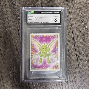 May include: A graded Pokémon card featuring Scyther from the Pokémon Merlin (1998) series. The card is encased in a clear protective holder and is graded NM/MINT 8. The card art depicts a green insectoid creature with large wings and a purple floral background.