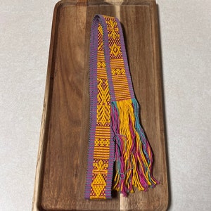May include: A woven yellow, pink, and purple belt with fringe on the ends. The belt is on a wooden surface.