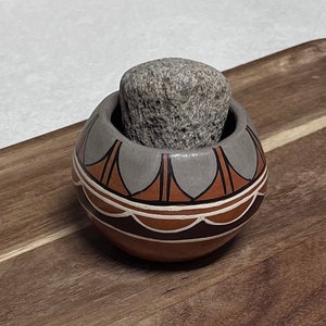 May include: A small, round, ceramic bowl with a geometric pattern in shades of brown, orange, and grey. A grey stone sits inside the bowl.