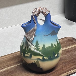 Peut inclure: Un petit vase en céramique avec une scène de montagne bleue, verte et brune. Le vase a une poignée en corde tressée et un accent de plume.