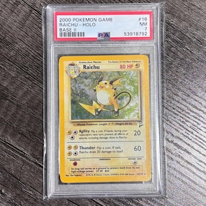 Puede incluir: Una carta Pokémon de Raichu de 2000, Base II, calificada como NM 7 y protegida en un estuche. La ilustración muestra a Raichu amarillo. La carta está marcada con el número #16.