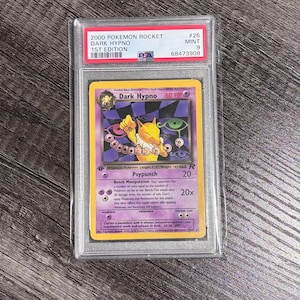 Könnte beinhalten: Eine bewertete Pokémon-Karte mit Dark Hypno aus dem 2000 Pokémon Rocket 1st Edition-Set. Die Karte ist in einem Schutzhalter eingeschlossen und wird als Mint 9 bewertet. Die Karte hat ein gelbes und lila Design mit dem Text "Dark Hypno".
