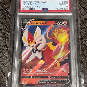 Puede incluir: Una carta Pokémon graduada con Cinderace V del set Fusión Strike. La carta muestra una colorida ilustración de Cinderace en acción, con texto que indica sus HP, ataque y otros detalles del juego. La carta está dentro de un soporte protector.