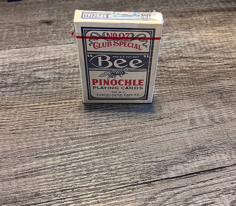 Puede incluir: Una baraja de cartas Bee en una caja azul y blanca. La caja dice "No. 07 Club Special" y "Bee" con una ilustraci&oacute;n de abeja. La caja tambi&eacute;n dice "Pinochle Playing Cards" y "The N.Y. Consolidated Card Co."