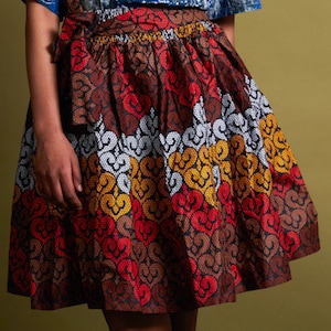 May include: A brown, red, white and yellow patterned skirt with a gathered waist. The skirt is made of a thick fabric and has a flared silhouette.