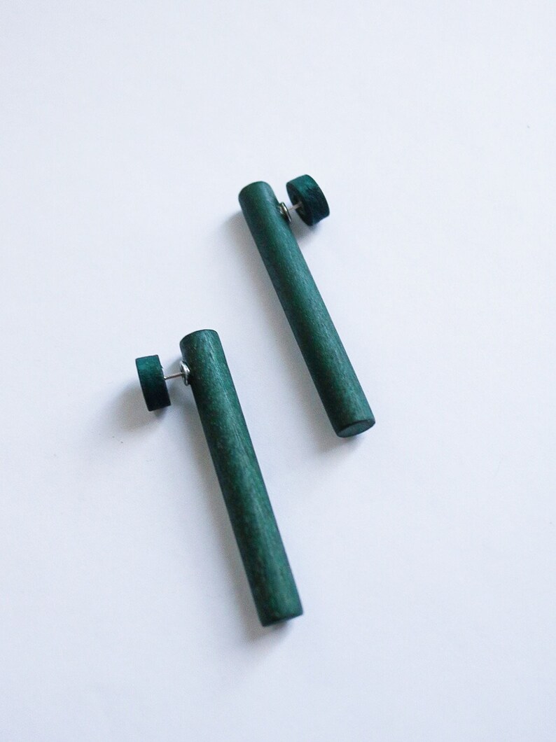 May include: A pair of dark green wooden earrings. Each earring is a long, thin cylinder with a small round disc at the top.