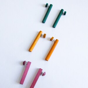 May include: Three pairs of wooden earrings in different colors. The first pair is teal green, the second pair is yellow, and the third pair is a deep pink.