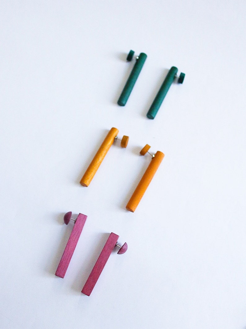 May include: Three pairs of wooden earrings in different colors. The first pair is teal green, the second pair is yellow, and the third pair is a deep pink.