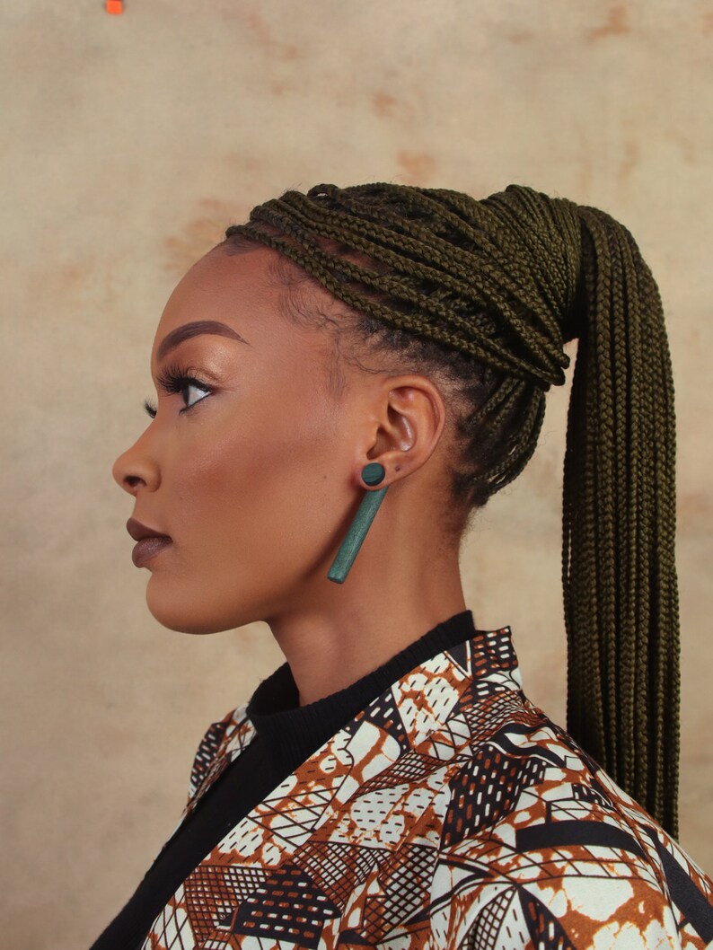 May include: A woman with dark skin is wearing a black turtleneck shirt and a brown and white patterned jacket. She has a high ponytail of dark green box braids. She is wearing a pair of long, dark green earrings.