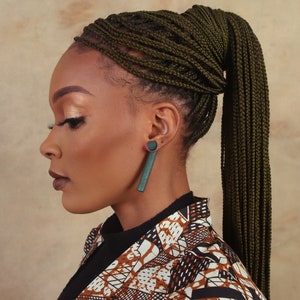 May include: A woman with dark skin is wearing a brown and white patterned jacket. Her hair is styled in a high ponytail with dark green braids. She is wearing a pair of dark green rectangular earrings.