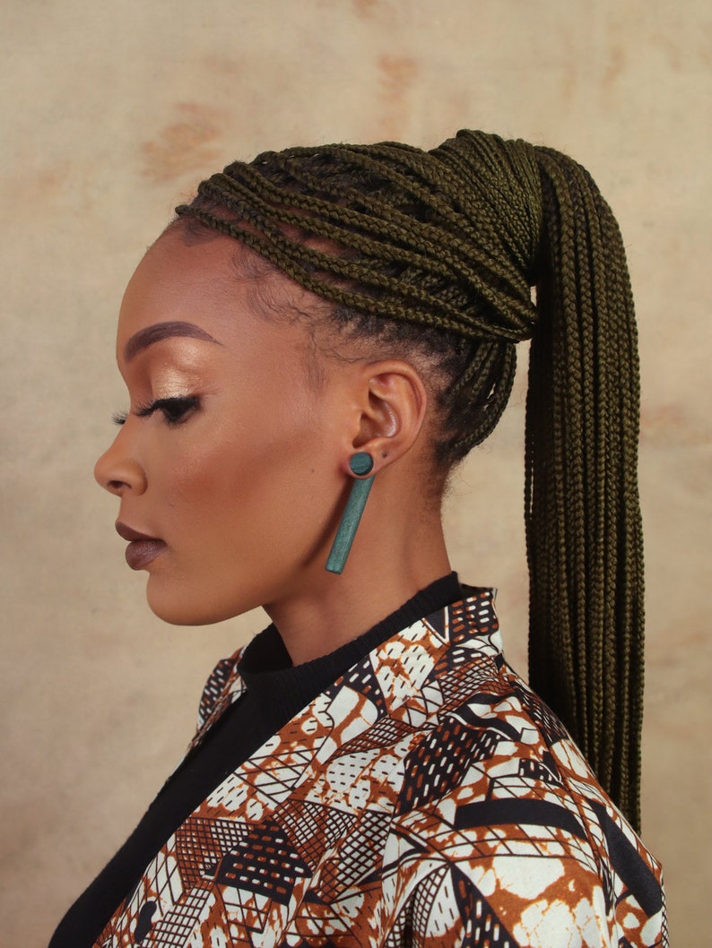 May include: A woman with dark skin is wearing a brown and white patterned jacket. Her hair is styled in a high ponytail with dark green braids. She is wearing a pair of dark green rectangular earrings.