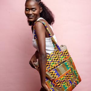 May include: A patterned tote bag with an African print design. The bag displays a patchwork of geometric shapes in yellow, blue, green, and red. It has a zipped pocket and strong straps. The background is a plain pink.