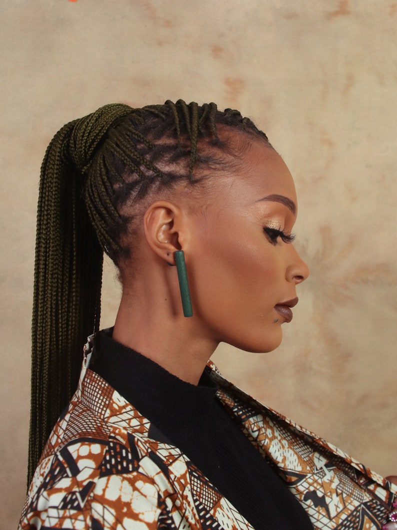 May include: A woman with dark skin is wearing a black turtleneck shirt and a brown and white patterned jacket. She has long green braids pulled back into a ponytail. She is wearing a pair of long, dark green earrings.
