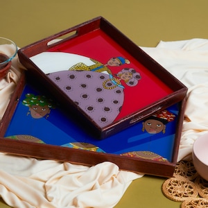 May include: Two wooden serving trays with colorful painted designs. One tray has a red background with a couple in traditional attire. The other tray has a blue background with a woman's face. A glass bowl and a pink bowl are also in the image.