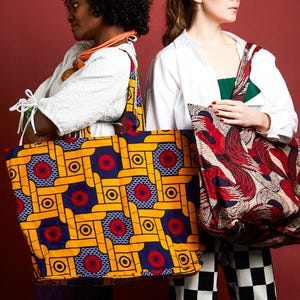 May include: Two tote bags with colorful geometric patterns. One bag features a yellow, blue, and red design, while the other has a red, black, and cream pattern. The bags are held by two people against a red background.