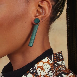May include: A long, dark green wooden earring with a round, dark green wooden top. The earring is worn in the left ear.