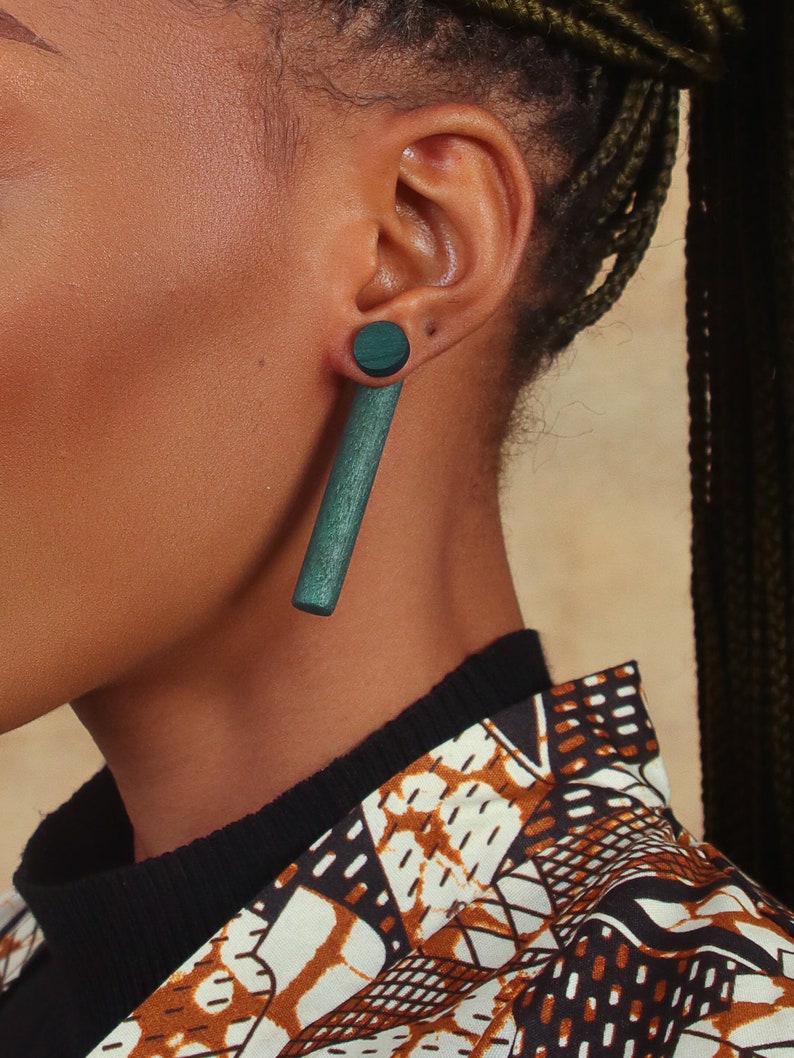 May include: A long, dark green wooden earring with a round, dark green wooden top. The earring is worn in the left ear.