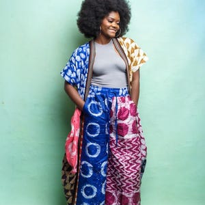 May include: A woman wearing a gray tank top, a colorful patterned kimono, and wide-leg pants. The kimono features blue, pink, and yellow patterns. The pants are blue and pink with geometric designs. The woman is smiling and wearing gold hoop earrings.