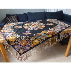 May include: A black rectangular tablecloth featuring intricate floral embroidery in shades of yellow, orange, purple, and white. The fabric is adorned with a central circular motif and decorative borders, finished with long, light-colored fringe trim.