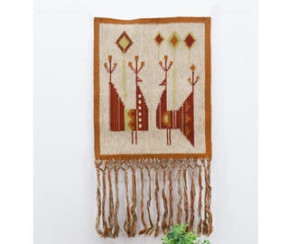 May include: A woven wall hanging with a beige background and a geometric design of stylized birds in shades of orange, brown, and yellow. The hanging has a fringe border.