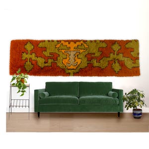 May include: A long, rectangular, orange and green shag rug with a central geometric design is displayed on a white wall. A green velvet sofa with black legs sits below the rug. A small plant stand and potted plants are also in the image.