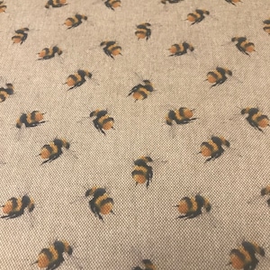 May include: A close-up shot of a beige fabric featuring a repeating pattern of bumblebees. The bees are illustrated in black and yellow, with small wings. The fabric has a woven texture, and the bees are evenly spaced.