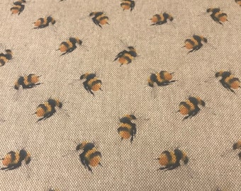 May include: Close-up of a beige fabric with a repeating pattern of bumblebees. The bees are depicted in black and yellow, with small wings. The fabric has a woven texture, and the bees are evenly spaced across the surface.