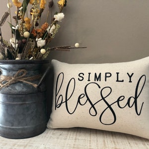 May include: Cream-colored rectangular pillow with the words "SIMPLY blessed" in black script. A galvanized metal milk jug with a twine bow holds dried flowers. The arrangement is on a white wooden surface.