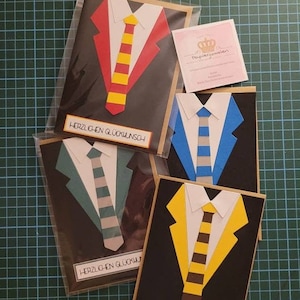 Könnte beinhalten: Vier handgemachte Grußkarten mit einem Anzug- und Krawatten-Design. Jede Karte hat einen Anzug und eine Krawatte in einer anderen Farbe. Die Karten sind schwarz mit weißem Text, der "HERZLICHEN GLÜCKWUNSCH" lautet.