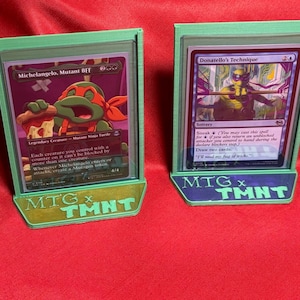 May include: Four green card holders with MTG x TMNT text, each holding a Magic: The Gathering card. The cards feature various illustrations and text. The holders are on a red surface.