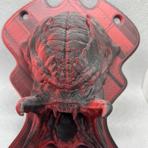 Puede incluir: Un busto de cabeza de Predator impreso en 3D, pintado en un esquema de colores negro y rojo. El busto está montado en una placa roja y negra con dos agujeros para colgar.