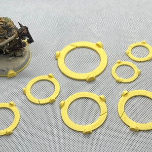 May include: A set of seven yellow plastic rings, some larger than others, used for gaming miniatures. The rings are designed to be placed around the base of a miniature to create a visual barrier.