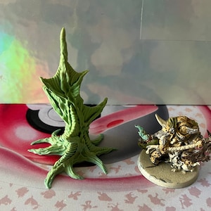 May include: A green, detailed, plastic, miniature tree with a base and a single, tall, branch. The tree is on a white and pink surface with a miniature, painted, plastic, warrior figure holding a weapon.
