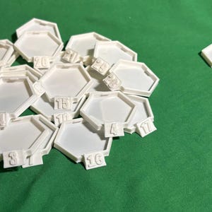 May include: A collection of white hexagonal plastic game tiles, each featuring a raised number on a small tab at the base. The set includes a large cluster of numbered pieces and a smaller group of separate, unnumbered hexagonal tiles.
