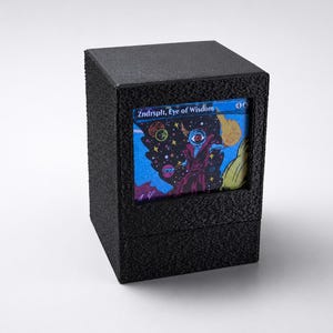 May include: A black, textured card storage box with a framed display of a fantasy art card. The card features a cosmic illustration with the text "Zndrsplt, Eye of Wisdom". The box is square and has a matte finish.