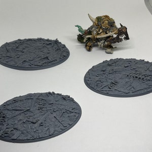 May include: Three gray, round, textured bases for miniature figures. The bases have a detailed, rough surface resembling a battlefield or a ruined city.