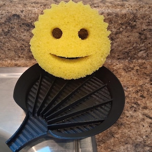 May include: A yellow smiley face sponge sits atop a black, fan-shaped sponge holder. The sponge has two dark brown eye holes and a curved mouth. The holder has a handle and is placed near a stainless steel sink.